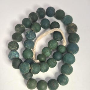 Könnte beinhalten: Eine Halskette aus runden, teal-farbenen Perlen, aufgereiht auf einer cremefarbenen Kordel. Die Perlen variieren in der Schattierung, einige weisen ein gesprenkeltes Aussehen auf. Die Halskette wird vor einem weißen Hintergrund präsentiert.