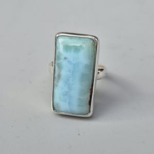 May include: A rectangular ring featuring a large, light blue gemstone with a silver band. The gemstone has a marbled appearance with shades of blue and white. The ring is set in a simple, polished silver setting.