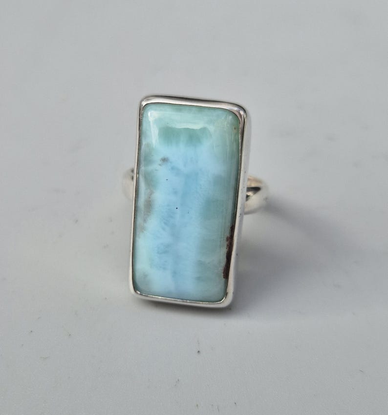 May include: A rectangular ring featuring a large, light blue gemstone with a silver band. The gemstone has a marbled appearance with shades of blue and white. The ring is set in a simple, polished silver setting.