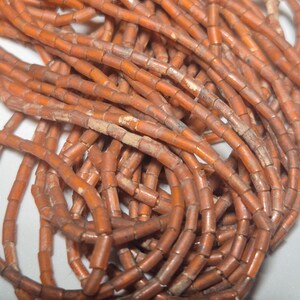 May include: Close-up of a collection of orange-brown cylindrical beads. The beads are strung together, forming multiple strands. The beads have a slightly weathered appearance, suggesting age or a vintage aesthetic. Ideal for jewelry making or craft projects.