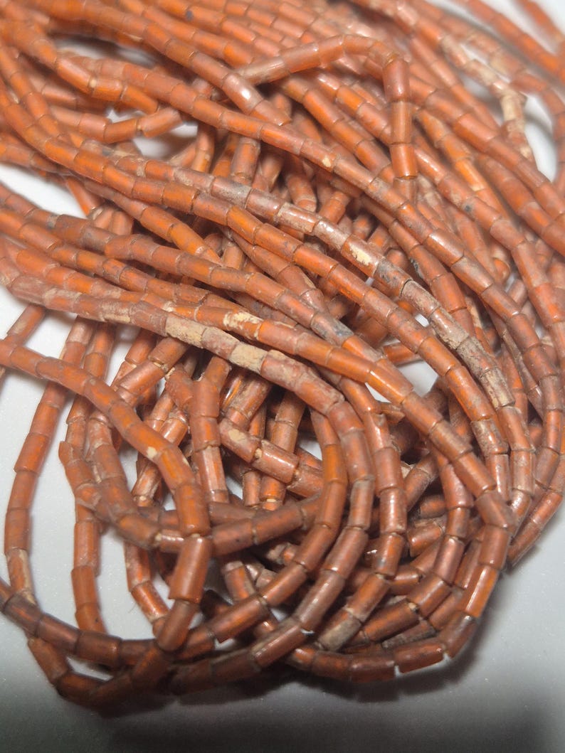 May include: Close-up of a collection of orange-brown cylindrical beads. The beads are strung together, forming multiple strands. The beads have a slightly weathered appearance, suggesting age or a vintage aesthetic. Ideal for jewelry making or craft projects.
