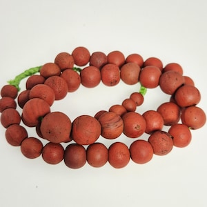 May include: A necklace of reddish-brown beads, varying in size, strung on a green cord. The beads have a matte finish and appear to be made of a natural material. The necklace is displayed against a white background.