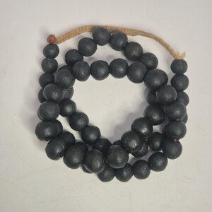 Könnte beinhalten: Eine Halskette aus schwarzen Holzperlen, auf einer naturfarbenen Kordel aufgereiht. Die Perlen sind rund und haben eine matte Oberfläche. Am Ende der Kordel befindet sich eine kleine rote Perle. Die Perlen sind kreisförmig angeordnet.