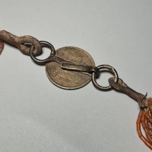 May include: Close-up of an antique necklace clasp. The clasp features a silver coin with engraved text, connected to a series of orange beads. The clasp is made of metal and leather, with a worn, vintage appearance.