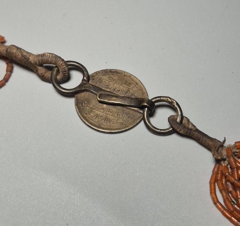 May include: Close-up of an antique necklace clasp. The clasp features a silver coin with engraved text, connected to a series of orange beads. The clasp is made of metal and leather, with a worn, vintage appearance.