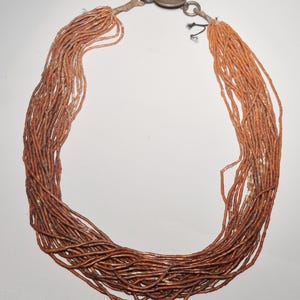 May include: A multi-strand necklace featuring numerous strands of small, reddish-brown beads. The necklace has a silver-colored clasp and connecting hardware. The beads are tightly strung, creating a full, textured appearance. The necklace is designed to be worn around the neck.