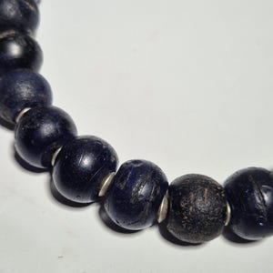 May include: Close-up of a dark blue beaded necklace. The beads are round and appear to be made of glass or stone, with a few showing a darker, almost black color. Silver-colored spacers are between the beads.