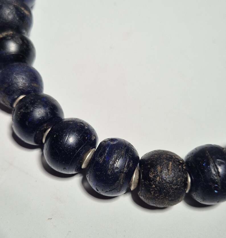 May include: Close-up of a dark blue beaded necklace. The beads are round and appear to be made of glass or stone, with a few showing a darker, almost black color. Silver-colored spacers are between the beads.