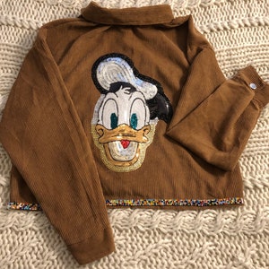 May include: A brown corduroy jacket with a sequined Donald Duck design on the front. The jacket has a beaded trim around the bottom.
