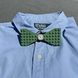Könnte beinhalten: Eine grüne Lego-Fliege mit einem kleinen Zifferblatt in der Mitte. Die Fliege befindet sich auf einem blauen Hemd mit Knöpfen und dem Logo von Polo by Ralph Lauren.