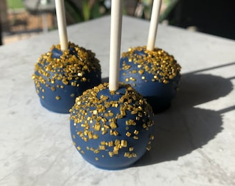 May include: Three blue cake pops with gold sprinkles on top. The cake pops are on white sticks and are arranged in a row on a white marble surface.
