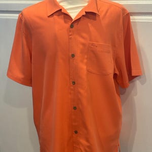 Pu&ograve; includere: Camicia arancione brillante a maniche corte con bottoni e una tasca sul petto. La camicia ha un tessuto strutturato e un colletto classico. Un piccolo logo &egrave; visibile sulla tasca. La camicia &egrave; esposta su un manichino.