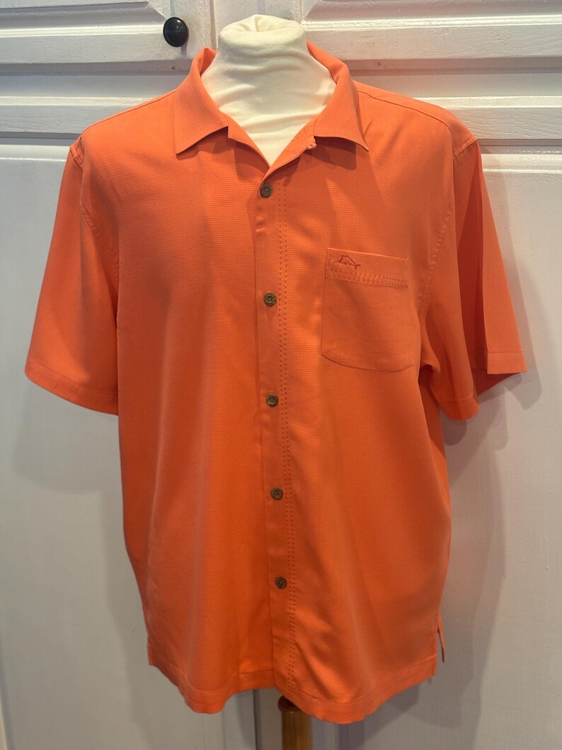 Pu&ograve; includere: Camicia arancione brillante a maniche corte con bottoni e una tasca sul petto. La camicia ha un tessuto strutturato e un colletto classico. Un piccolo logo &egrave; visibile sulla tasca. La camicia &egrave; esposta su un manichino.