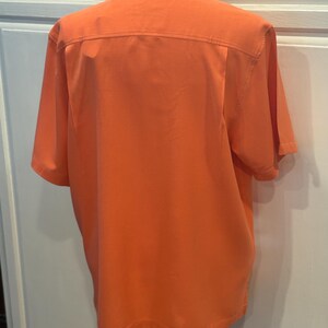 Pu&ograve; includere: Camicia arancione a maniche corte, vista da dietro. La camicia ha un colletto e un pannello strutturato sulla parte superiore della schiena. Il tessuto sembra essere un materiale leggero e traspirante. La camicia &egrave; su un manichino.