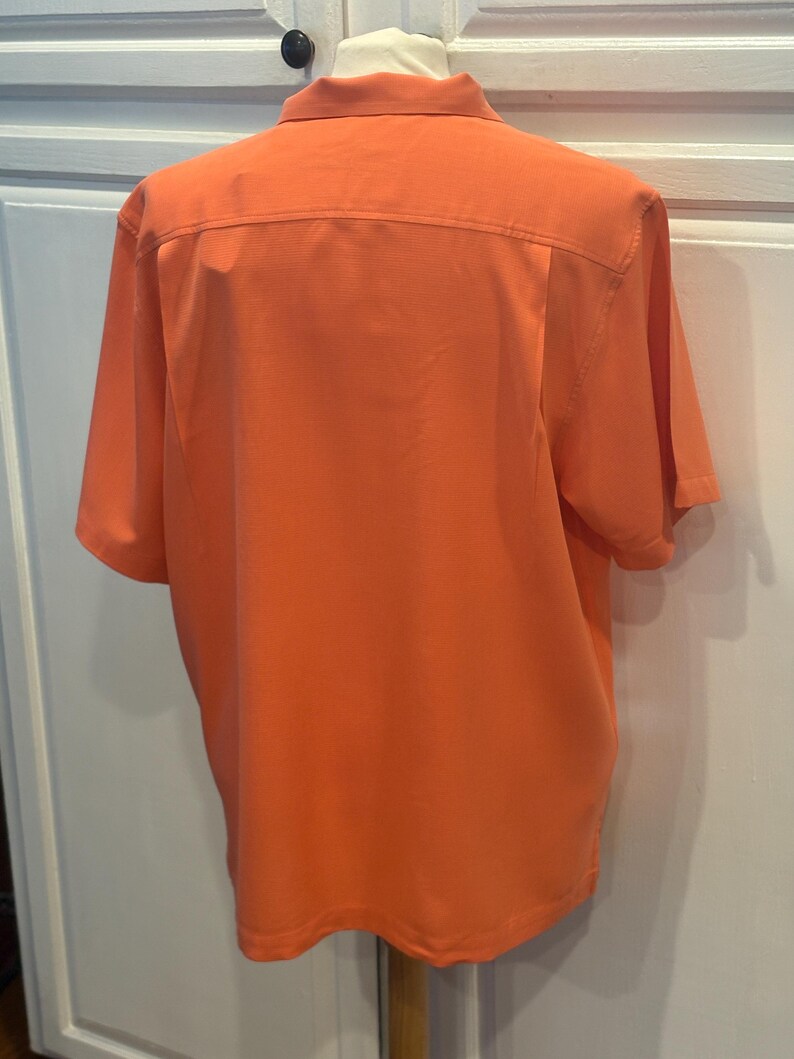 Pu&ograve; includere: Camicia arancione a maniche corte, vista da dietro. La camicia ha un colletto e un pannello strutturato sulla parte superiore della schiena. Il tessuto sembra essere un materiale leggero e traspirante. La camicia &egrave; su un manichino.