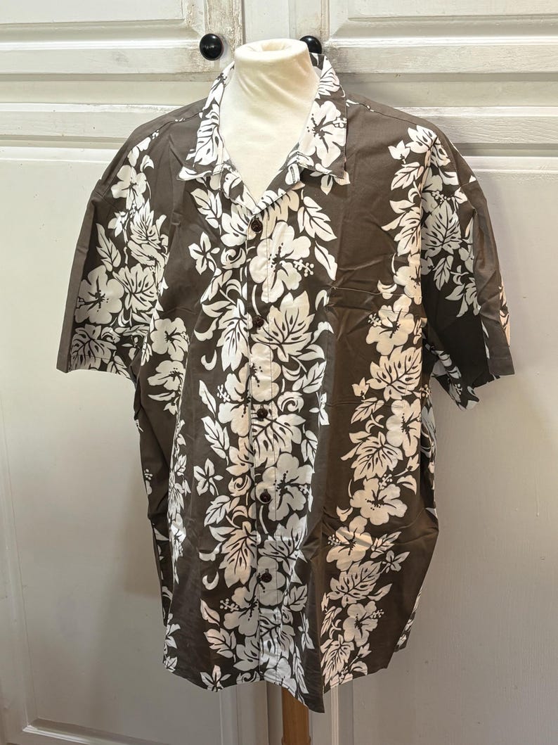 May include: A brown short-sleeved button-up shirt with a white floral print. The shirt has a classic collar and a button-up front.