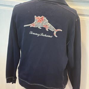 May include: A navy blue sweatshirt with a decorative fish graphic on the back, featuring red and white floral accents. The sweatshirt has a zip-up collar and the "Tommy Bahama" logo is printed below the graphic.
