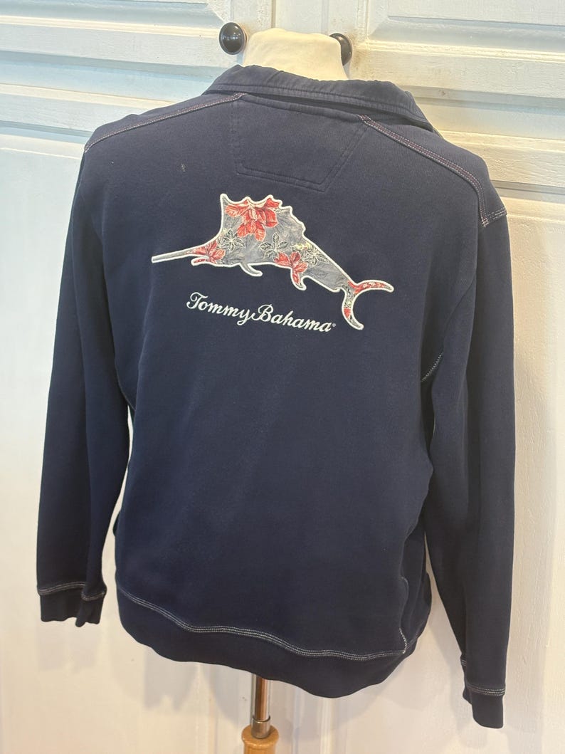 May include: A navy blue sweatshirt with a decorative fish graphic on the back, featuring red and white floral accents. The sweatshirt has a zip-up collar and the "Tommy Bahama" logo is printed below the graphic.