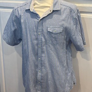 Könnte beinhalten: Ein kurzärmeliges Hemd mit blau-weißem Vichy-Karo-Muster. Das Hemd hat eine einzelne Brusttasche und einen kontrastierenden weißen Kragen. Das Hemd wird auf einer Schaufensterpuppe präsentiert.