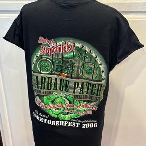 May include: Black t-shirt featuring a green motorcycle graphic within a chain design. The shirt includes text that reads "Cabbage Patch", "Biker Bar", and "Biketoberfest 2006". The shirt is displayed on a mannequin.