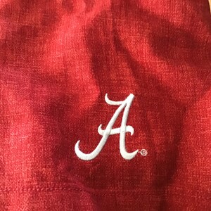 May include: Close-up of a red fabric with a white embroidered letter "A". The fabric has a textured appearance. The letter "A" is in a cursive style and is likely part of a team or school logo.