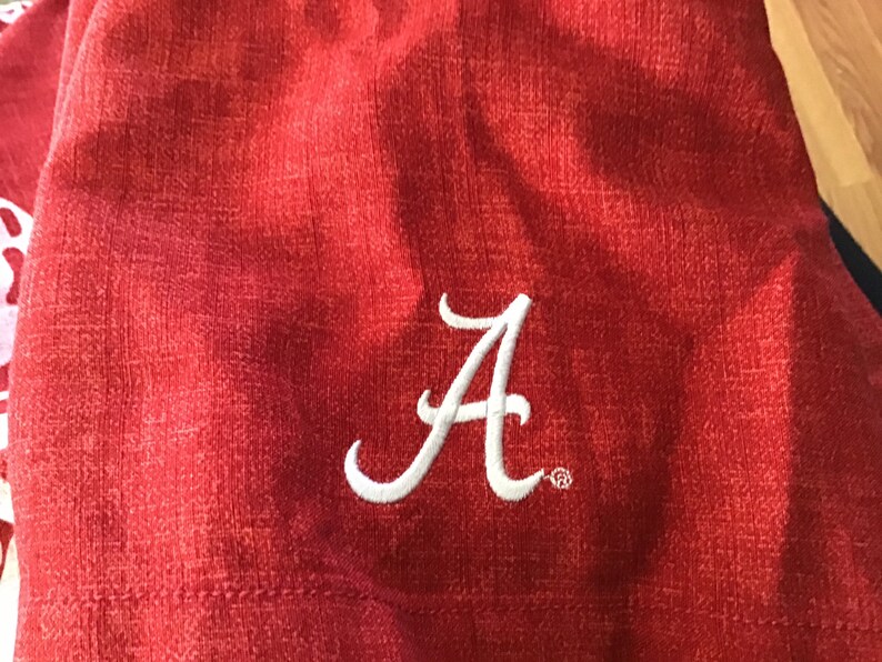May include: Close-up of a red fabric with a white embroidered letter "A". The fabric has a textured appearance. The letter "A" is in a cursive style and is likely part of a team or school logo.