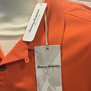 Pu&ograve; includere: Primo piano di una camicia arancione con bottoni e tessuto strutturato. La camicia ha una tasca e un'etichetta con il logo "Tommy Bahama" e il design a foglie. La camicia &egrave; esposta su un manichino.