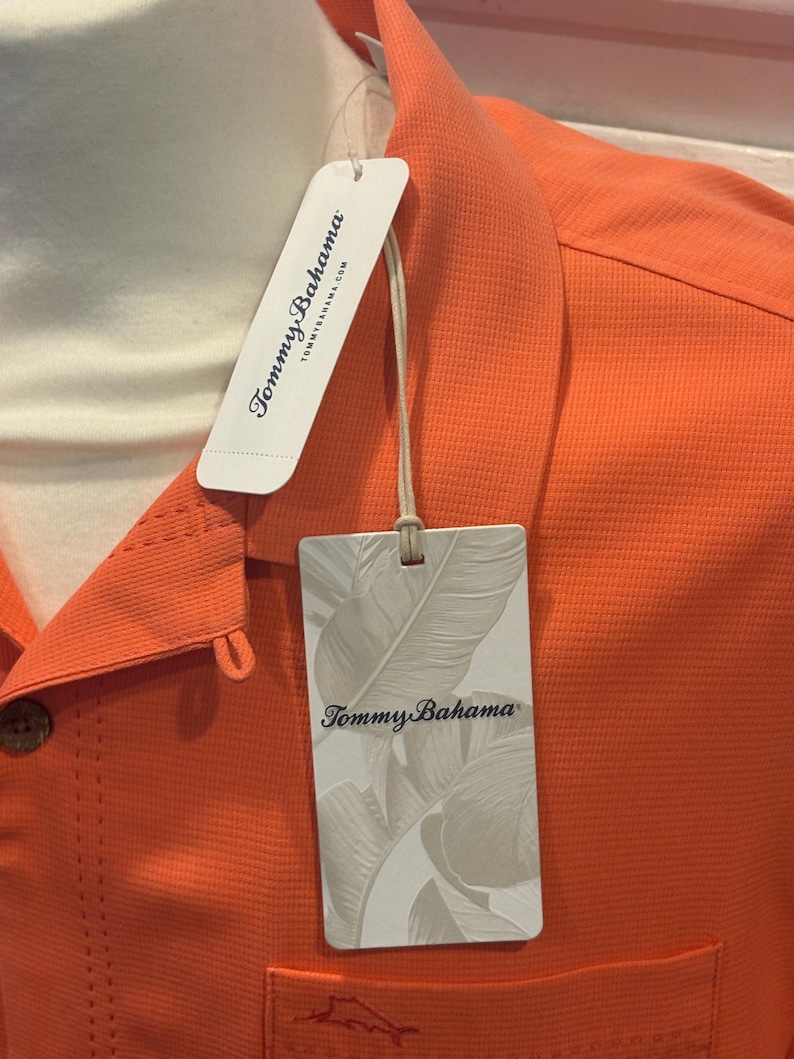 Pu&ograve; includere: Primo piano di una camicia arancione con bottoni e tessuto strutturato. La camicia ha una tasca e un'etichetta con il logo "Tommy Bahama" e il design a foglie. La camicia &egrave; esposta su un manichino.