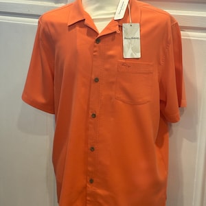Pu&ograve; includere: Camicia arancione brillante a maniche corte con bottoni e una tasca sul petto. La camicia ha un colletto classico e un'etichetta con la scritta "Tommy Bahama". La camicia &egrave; esposta su un manichino.