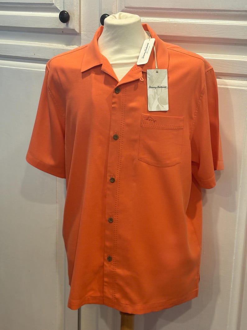 Pu&ograve; includere: Camicia arancione brillante a maniche corte con bottoni e una tasca sul petto. La camicia ha un colletto classico e un'etichetta con la scritta "Tommy Bahama". La camicia &egrave; esposta su un manichino.