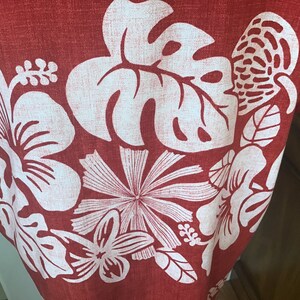 May include: Close-up of a red fabric with a white floral pattern. The design features large hibiscus flowers, monstera leaves, and other tropical elements. The fabric has a textured appearance.