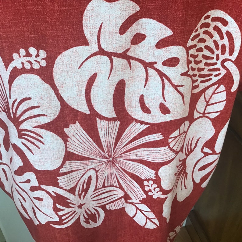 May include: Close-up of a red fabric with a white floral pattern. The design features large hibiscus flowers, monstera leaves, and other tropical elements. The fabric has a textured appearance.