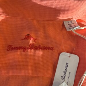 Pu&ograve; includere: Polo color corallo con trama strutturata. Presenta un logo marlin rosso ricamato e il nome del marchio "Tommy Bahama" in corsivo rosso. &Egrave; presente un'etichetta bianca con il nome del marchio "Bahama".