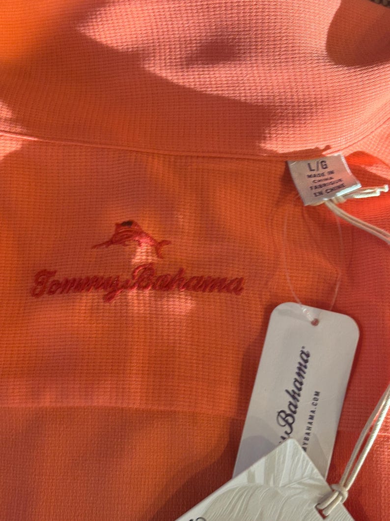 Pu&ograve; includere: Polo color corallo con trama strutturata. Presenta un logo marlin rosso ricamato e il nome del marchio "Tommy Bahama" in corsivo rosso. &Egrave; presente un'etichetta bianca con il nome del marchio "Bahama".