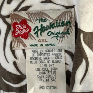 May include: A white tag with brown trim and black text. The tag reads "The Hawaiian Original 4XL Made in Hawaii" and includes care instructions. The tag is attached to a white shirt with a brown floral pattern.