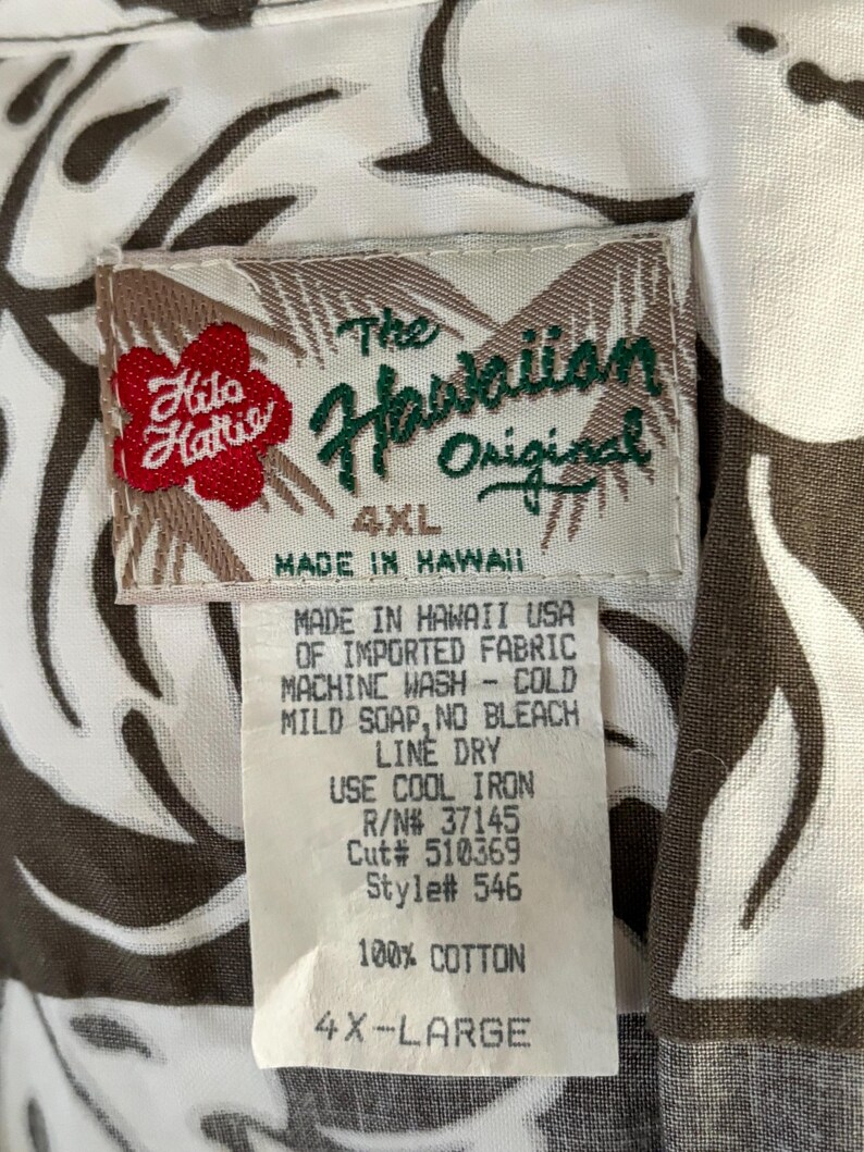 May include: A white tag with brown trim and black text. The tag reads "The Hawaiian Original 4XL Made in Hawaii" and includes care instructions. The tag is attached to a white shirt with a brown floral pattern.