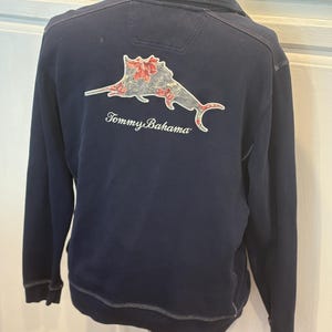 May include: Navy blue long-sleeve sweatshirt with a sailfish graphic on the back, featuring red and white floral accents. The text "Tommy Bahama" is below the graphic. The sweatshirt has a zip-up collar and is displayed on a mannequin.