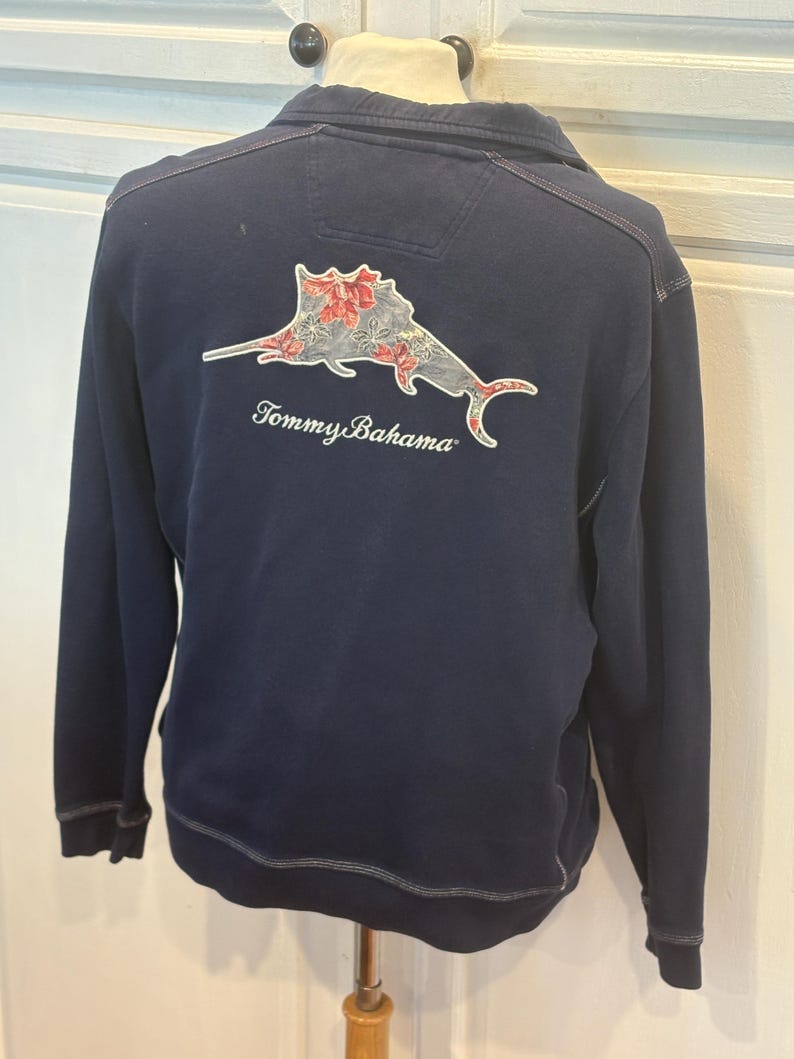 May include: Navy blue long-sleeve sweatshirt with a sailfish graphic on the back, featuring red and white floral accents. The text "Tommy Bahama" is below the graphic. The sweatshirt has a zip-up collar and is displayed on a mannequin.