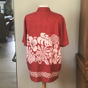 May include: A red short-sleeved shirt with a white floral design across the lower portion. The shirt is displayed on a mannequin. The floral pattern includes hibiscus flowers, monstera leaves, and other tropical elements. The bottom hem features a geometric pattern.