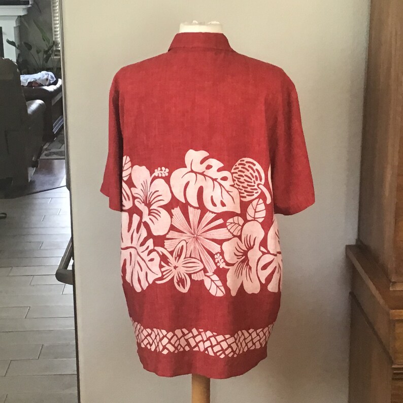 May include: A red short-sleeved shirt with a white floral design across the lower portion. The shirt is displayed on a mannequin. The floral pattern includes hibiscus flowers, monstera leaves, and other tropical elements. The bottom hem features a geometric pattern.