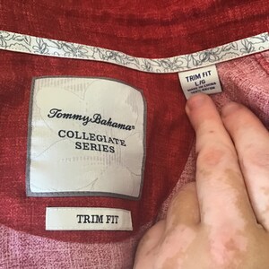 May include: Close-up of a red Tommy Bahama Collegiate Series shirt. The shirt has a floral pattern on the collar and the label reads "TRIM FIT L/G MADE IN CHINA 100% RAYON".