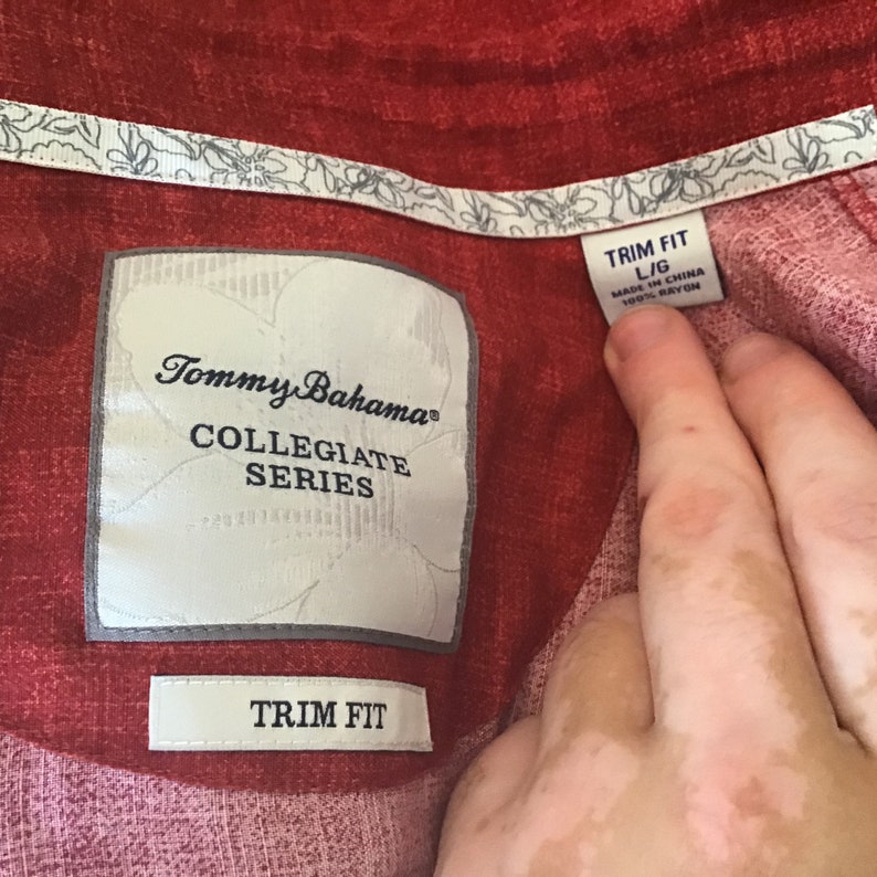 May include: Close-up of a red Tommy Bahama Collegiate Series shirt. The shirt has a floral pattern on the collar and the label reads "TRIM FIT L/G MADE IN CHINA 100% RAYON".