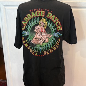 Könnte beinhalten: Schwarzes T-Shirt mit einem grafischen Design. Das Design enthält den Text "Sopotnick's Cabbage Patch" in Grün und Rot sowie eine Illustration zum Thema Wrestling. Die Worte "Samsula, Florida 2004" sind ebenfalls vorhanden.