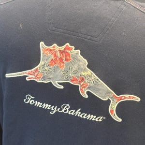 May include: Navy blue polo shirt featuring a floral marlin design on the back. The marlin is grey with red and white floral accents. The words "Tommy Bahama" are embroidered in white.