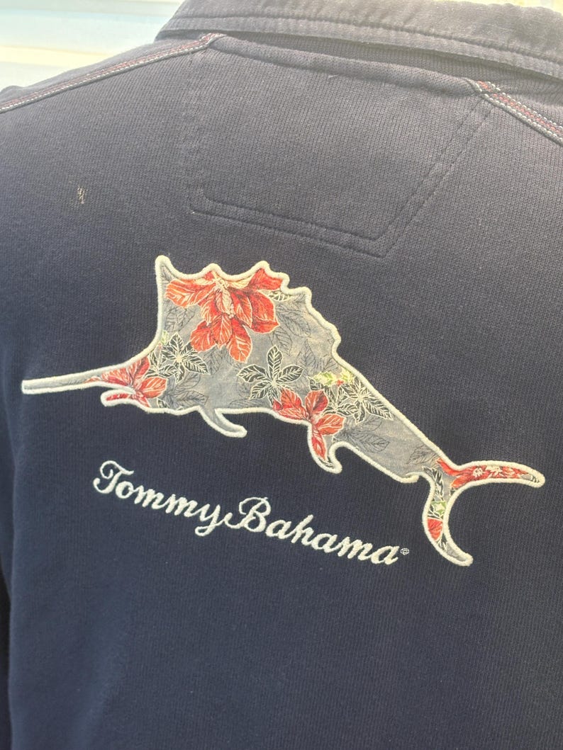 May include: Navy blue polo shirt featuring a floral marlin design on the back. The marlin is grey with red and white floral accents. The words "Tommy Bahama" are embroidered in white.