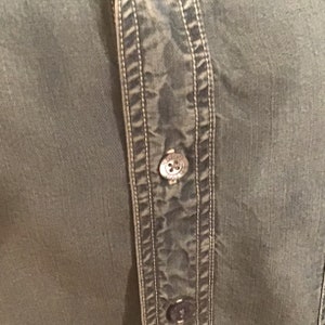 May include: Close-up of a green button-up shirt with a double row of stitching down the front. The shirt has a button with a logo on it.