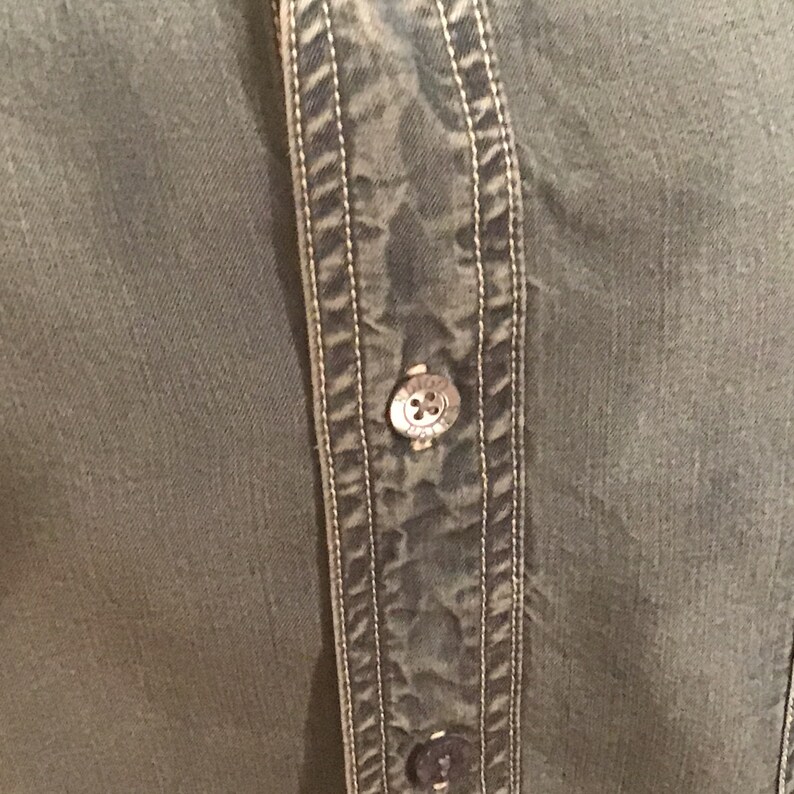May include: Close-up of a green button-up shirt with a double row of stitching down the front. The shirt has a button with a logo on it.