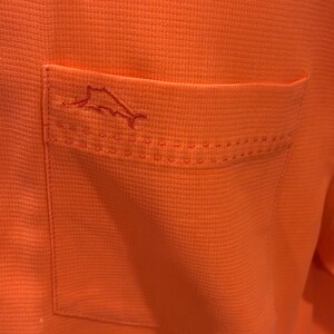 Pu&ograve; includere: Primo piano di una camicia arancione con trama strutturata. La camicia presenta una tasca sul lato sinistro con un contorno cucito e un piccolo logo di pesce ricamato.