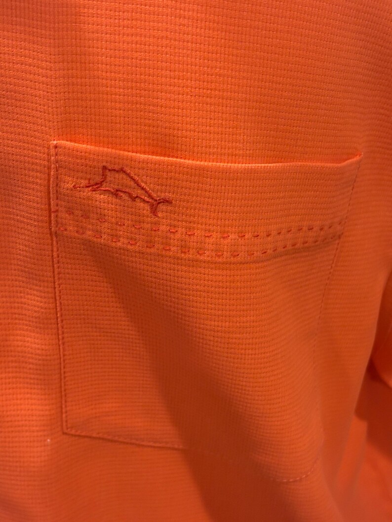 Pu&ograve; includere: Primo piano di una camicia arancione con trama strutturata. La camicia presenta una tasca sul lato sinistro con un contorno cucito e un piccolo logo di pesce ricamato.