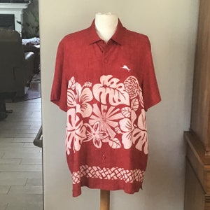 May include: Red short-sleeve button-down shirt with a white floral design. The shirt features a white logo of a fish on the upper left chest. The bottom hem has a white geometric pattern.
